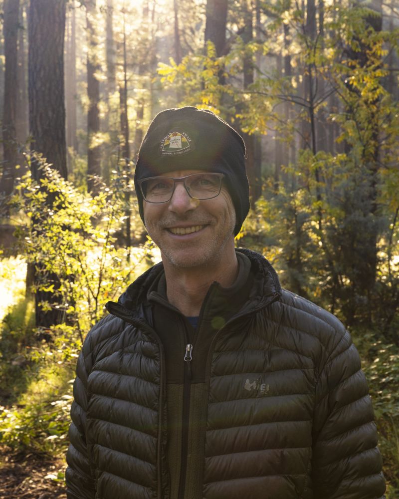 A man in glasses and a black jacket and a black knit cap standing in a sunlit forest