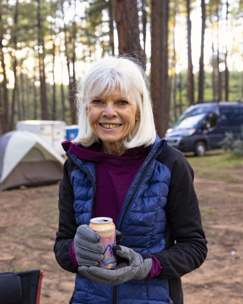 A woman with short white hair in a puffy jacket standing in a forested campsite